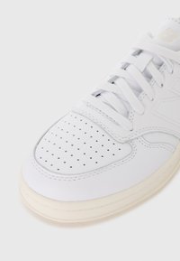 Valge nahkne tennis, millel on perforatsioonidega varvaskarp, lamedad valged paelad ja kreemjas kummitald. Omab peent logo keeles.