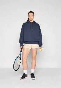 Felpa navy con lacci, pantaloni corti beige, scarpe da ginnastica nere, che tiene in mano una racchetta da tennis e una pallina da tennis gialla. Sfondo bianco.
