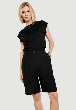 Blondynka w czarnej, bezrękawnikowej bluzce i czarnych bermudach z wysokim stanem, stojąca z jedną ręką w kieszeni.