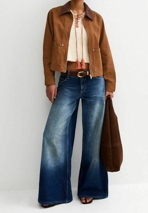Person iført brun jakke, cremefarvet snøre crop top, vidde ben falmede blå jeans, holder en brun taske.