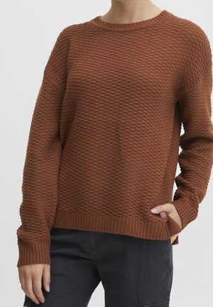 Person trägt einen strukturierten braunen Strickpullover mit langen Ärmeln und schwarze Hose, die Hand teilweise in der Tasche verstaut.