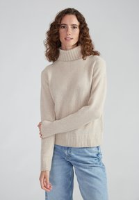 Beiger Rollkragenpullover aus weichem Material mit gerippten Bündchen und Saum, kombiniert mit hellblauen Jeans. Einfaches, gemütliches Design.