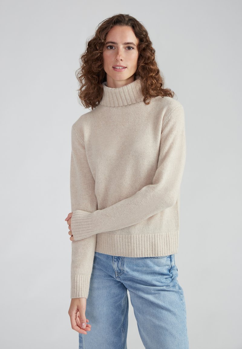 Beiger Rollkragenpullover aus weichem Material mit gerippten Bündchen und Saum, kombiniert mit hellblauen Jeans. Einfaches, gemütliches Design.