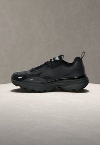 Baskets athlétiques noires avec semelle épaisse, fermeture à lacets à l'avant, et languettes à la langue et au talon, présentées sur une surface claire sur fond neutre.