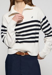 Femme portant un pull-over blanc à fermeture éclair avec des rayures horizontales noires, un pantalon bleu marine et une ceinture marron avec une boucle dorée.