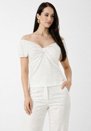 Donna con capelli lunghi e scuri che indossa una maglietta bianca scollata sulle spalle e pantaloni abbinati con motivo a occhielli, orecchini a cerchio dorati e braccialetto.