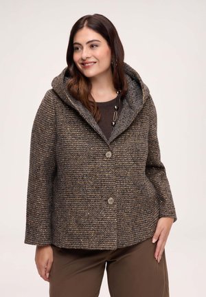 Woman wearing a textured brown buttoned coat with a wide collar hood and brown pants, smiling and looking to her right.