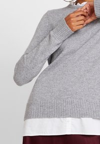 Grauer Strickpullover mit geripptem Saum und Bündchen, über einem weißen Hemd mit Kragen getragen. Weiche Textur mit einem schlichten Design und ohne sichtbare Muster.