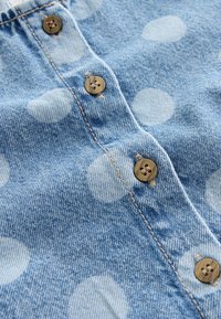 Denimstof i lyseblå med hvide polkaprikker. Har brune knapper og synlige syninger langs knappelisten.