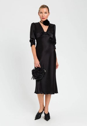 Donna in piedi con un abito midi in raso nero con le maniche arricciate, che tiene una borsa rotonda nera floreale e indossa décolleté nere a punta.
