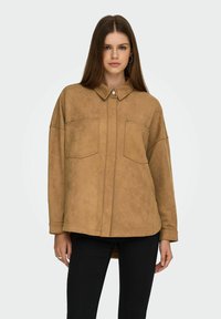 Bruine faux suede button-up shirt met twee borstzakken, een puntige kraag en een ontspannen pasvorm, gedragen over zwarte broek.