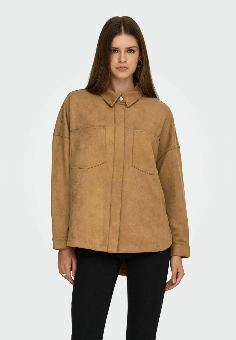 Bruine faux suede button-up shirt met twee borstzakken, een puntige kraag en een ontspannen pasvorm, gedragen over zwarte broek.