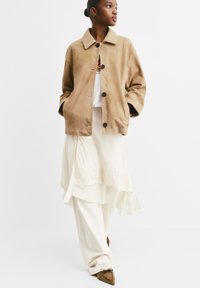 Giacca in suede beige con collo, caratterizzata da grandi bottoni e vestibilità rilassata, indossata sopra una gonna crema asimmetrica con bordo in pizzo.