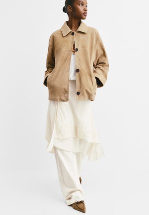 Giacca in suede beige con collo, caratterizzata da grandi bottoni e vestibilità rilassata, indossata sopra una gonna crema asimmetrica con bordo in pizzo.