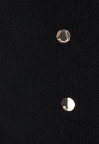 Schwarzer gestrickter Stoff mit zwei glänzenden goldenen Knöpfen, von denen jeder am Rand mit kleinem kursivem Text graviert ist.