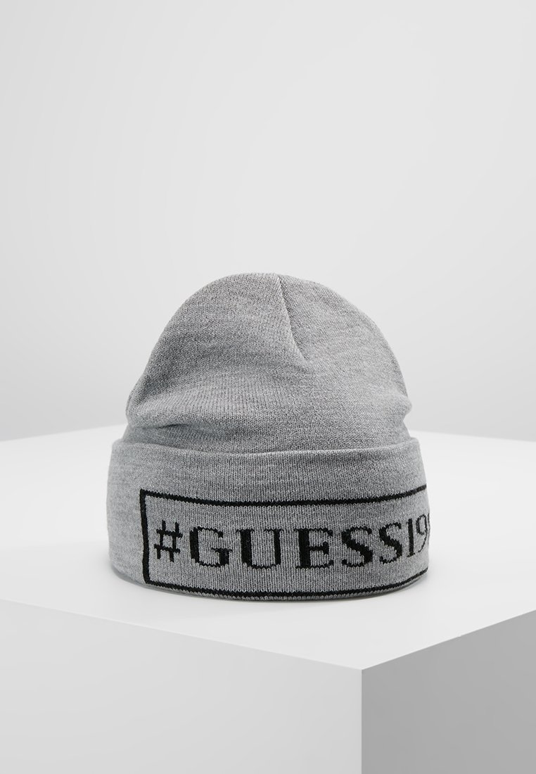 Bonnet en maille gris avec bord replié affichant "#GUESS1981" en texte noir dans un cadre rectangulaire, posé sur une surface blanche.