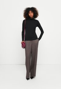 Frau mit lockigem Haar, die einen schwarzen Rollkragenpullover, braune weite Hosen, schwarze spitze Schuhe und eine burgunderfarbene Schultertasche trägt, steht vor einer weißen Wand.