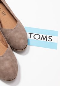 Chaussures slip-on perforées beiges à bouts ronds, accompagnées d'une étiquette TOMS bleu et blanc avec un texte noir en gras.