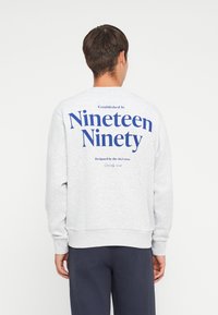 Sweatshirt gris à col ras du cou avec un grand imprim�é bleu au dos, portant les inscriptions "Établi en mille neuf cents quatre-vingt-dix" et "Designé par l'équipe J&J."