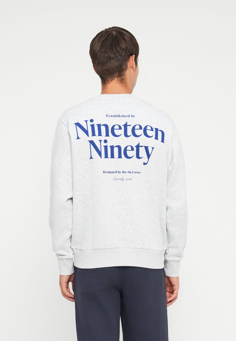 Sweatshirt gris à col ras du cou avec un grand imprimé bleu au dos, portant les inscriptions "Établi en mille neuf cents quatre-vingt-dix" et "Designé par l'équipe J&J."