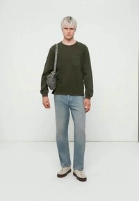 Ung person med kort blond hår iført en olivengrønn genser, lyseblå jeans, hvite joggesko og en grå skulderveske.