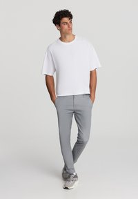 Mężczyzna w białej, oversize'owej koszulce, jasnoszarych, dopasowanych spodniach i beżowych sneakersach stoi z rękami w kieszeniach, patrząc w prawo na jednolitym tle.