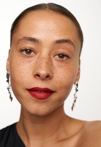 Närbild av en person med bruna ögon och fräknar, som bär djärv röd läppstift och silverfärgade, abstrakta örhängen mot en enkel vit bakgrund.