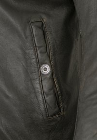 Schwarze Lederjacke mit einer Reißverschlusstasche und einem silbernen Druckknopfverschluss. Die Oberfläche ist glatt mit leichten Falten und einem matten Finish.