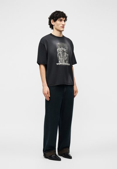 Νεαρός άνδρας που στέκεται φορώντας μαύρο υπερμεγέθες γραφικό t-shirt, φαρδιά σκούρα παντελόνια και μαύρα μυτερά παπούτσια, μπροστά σε απλό φωτεινό φόντο.