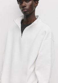 Witte zip-up sweatshirt met hoge kraag en relaxed fit. Heeft een zachte textuur en een zilveren rits. Gelaagd over een donker shirt.