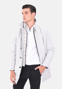 Giacca grigia chiara con un design minimalista, caratterizzata da un alto collo, chiusura con bottoni e un rivestimento interno trapuntato. Indossata su una camicia bianca.