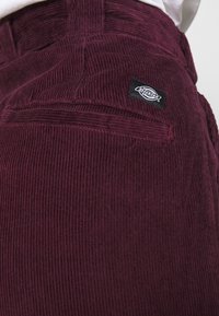 Pantalon en velours côtelé bordeaux avec textures verticales, doté d'une poche arrière et d'une étiquette noire Dickies cousue à la ceinture.