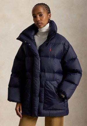 Donna che indossa una giacca imbottita oversize blu navy con collo alto, dolcevita bianco e pantaloni color cammello su sfondo semplice.