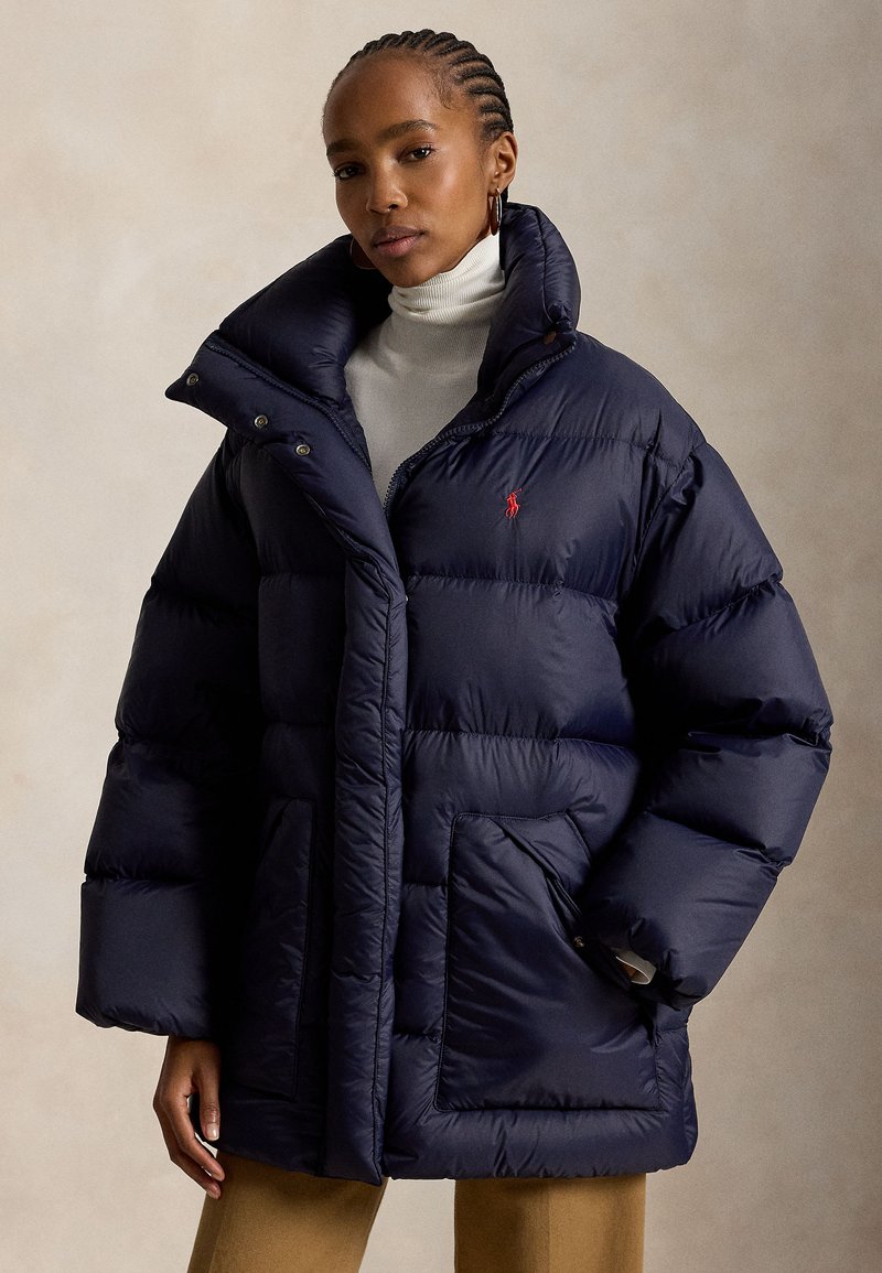 Femme portant une doudoune bleu marine oversize avec col montant, un col roulé blanc et un pantalon beige sur fond uni.