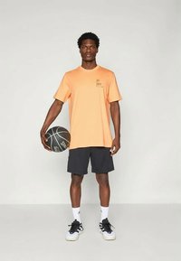 Πορτοκαλί αθλητικό μπλουζάκι με μαύρο λογότυπο, συνδυασμένο με μαύρες βερμούδες. Το άτομο κρατάει μια μαύρη μπάλα μπάσκετ και φοράει λευκά και μαύρα αθλητικά παπούτσια.