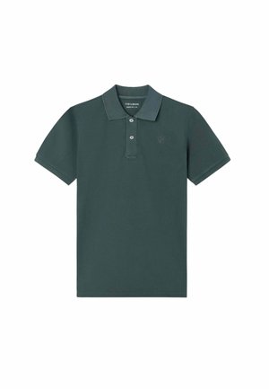 Hombre con camiseta polo verde oscuro, pantalones cortos beige, zapatos náuticos marrones y gafas de sol, de pie con una mano en el bolsillo.