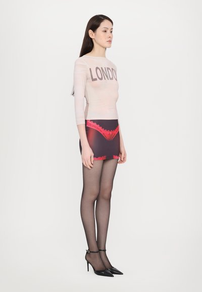 Durchsichtige Langarm-Bluse mit "LONDON"-Aufschrift, hoch taillierter Minirock mit roten Spitzenakzenten, schwarze durchsichtige Strumpfhosen und schwarze High-Heels.