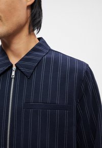 Άνδρας που φοράει ένα ναυτικό μπλε ζακέτα με φερμουάρ και λευκές κατακόρυφες ρίγες, με τσέπη στο στήθος, απεικονίζε ται από το λαιμό μέχρι το μέσο του κορμού.