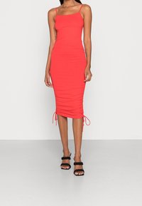 Robe rouge ajustée avec des bretelles spaghetti, des détails froncés et des liens ajustables à l'ourlet. Associée à des sandales noires à lanières.