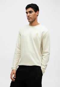 Εκρού φούτερ με μακριά μανίκια και στρογγυλή λαιμόκοψη, με κεντημένο χρυσό λογότυπο στο στήθος. Λείο ύφασμα, συνδυασμένο με μαύρο παντελόνι.