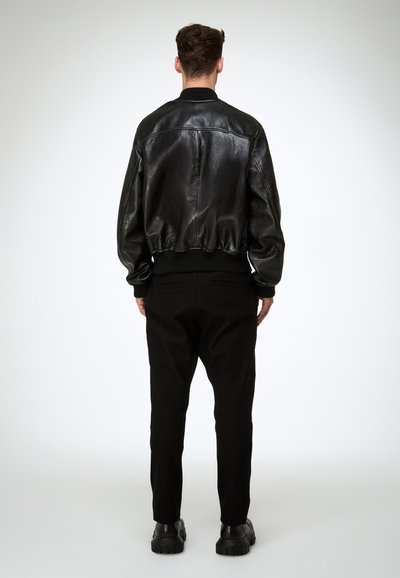Fényes fekete bőr bombázódzsekit, fekete nadrágot és fekete masszív cipőt viselő férfi hátulról, világos háttér előtt áll.