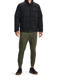 Czarna puchowa kurtka na suwak, oliwkowe spodnie dresowe z zakrzywionymi szwami oraz czarne buty sportowe z brązowymi akcentami.