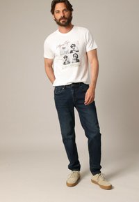 T-shirt en coton blanc avec un graphisme imprimé, associé à un jean en denim bleu foncé. Les chaussures sont en daim beige avec des lacets blancs et des semelles en caoutchouc.