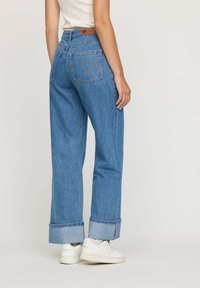 Personne portant un jean bleu avec revers et des baskets blanches se tenant contre un fond clair uni, de dos.