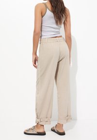 Pantalons larges beiges avec des ourlets retroussés, présentant des boutons au niveau de l'ourlet. Associés à un débardeur rayé gris et blanc et des sandales marron.