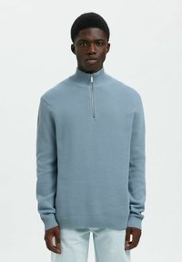 Mladic obučen u svijetloplavu pletenu majicu s kratkim patentnim zatvaračem i svijetlim hlačama, stoji naslonjen na jednostavnu bijelu pozadinu.