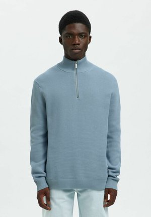 Un jeune homme portant un pull en tricot bleu clair avec une courte fermeture éclair et un pantalon de couleur claire, se tenant contre un fond blanc uni.