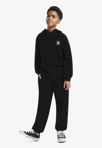 Σετ μαύρου φλις φούτερ με κουκούλα και παντελόνι φόρμας με μπροστινή τσέπη. Διακοσμημένο με πολύχρωμο λογότυπο αστεριού στο στήθος και μανσέτες στα πόδια του παντελονιού. Μαύρα αθλητικά παπούτσια.