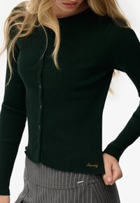 Ciemnozielony kardigan z prążkowanej tkaniny, z długim rękawem, okrągłym dekoltem, guziczkami z przodu oraz haftowanym logo na dolnym brzegu.