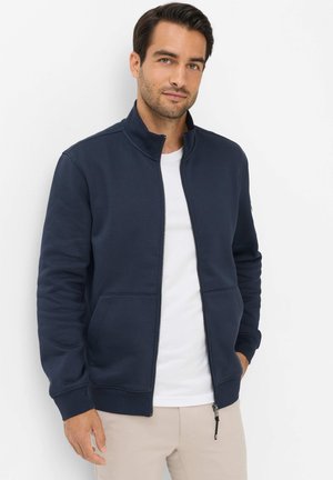 Mann trägt eine marineblaue Jacke mit Reißverschluss über einem weißen Hemd, steht mit einer Hand in der Tasche vor einem schlichten weißen Hintergrund.